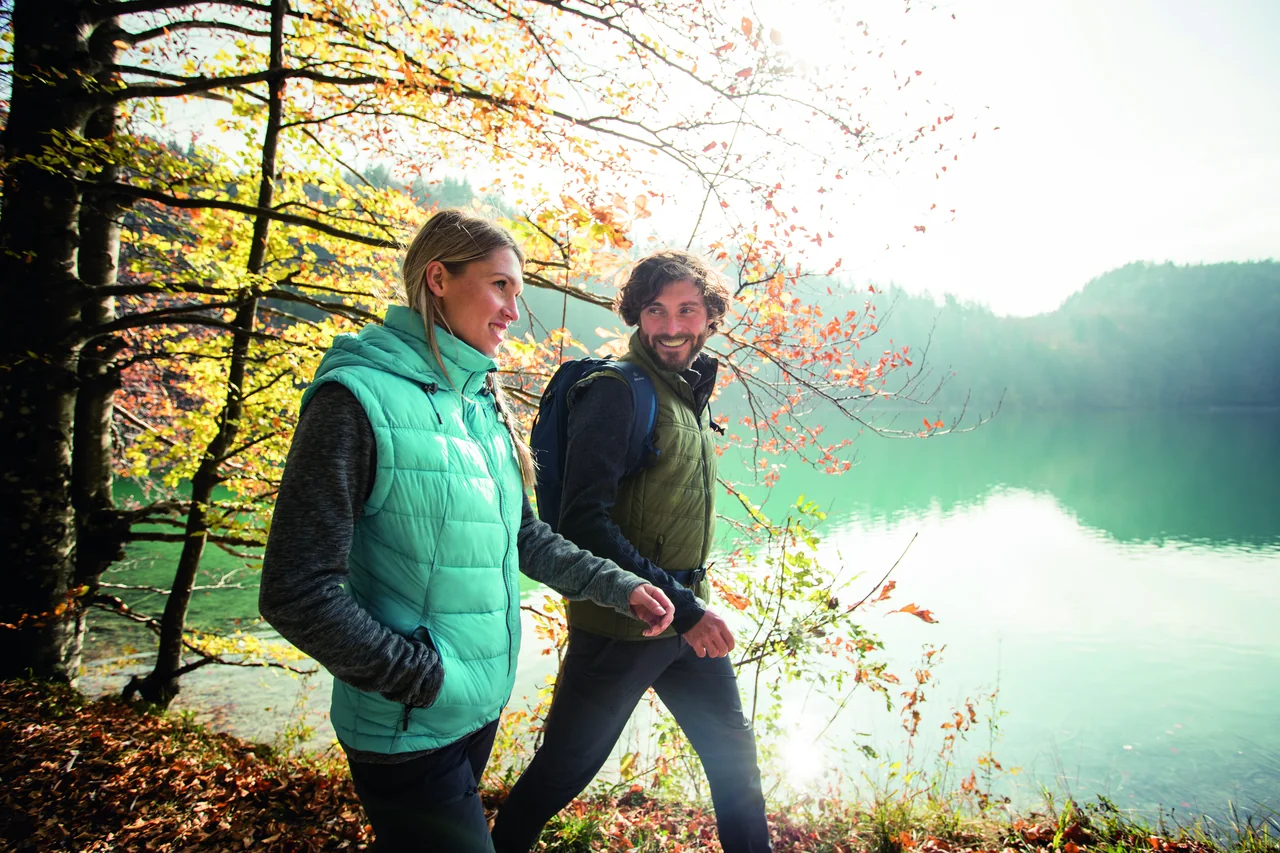 Zwei Wanderer in Outdoor-Bekleidung an einem See im herbstlichen Wald.